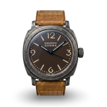 A Gruppo Gamma wristwatch with an aged steel dark gray case, a tropical dark brown dial with beige Arabic numerals and steel blue hands, and a brown leather strap.
